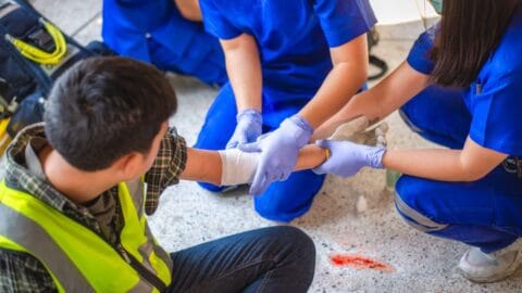 Emergency First Aid at Work online course: paramedics treating a worker in a hi-vis vest on the ground